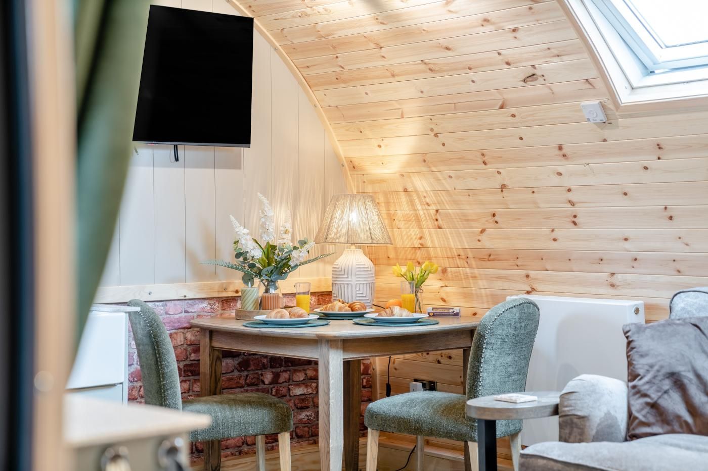 A cozy dining area with a wooden ceiling, a table set with plates, a lamp, and flowers.