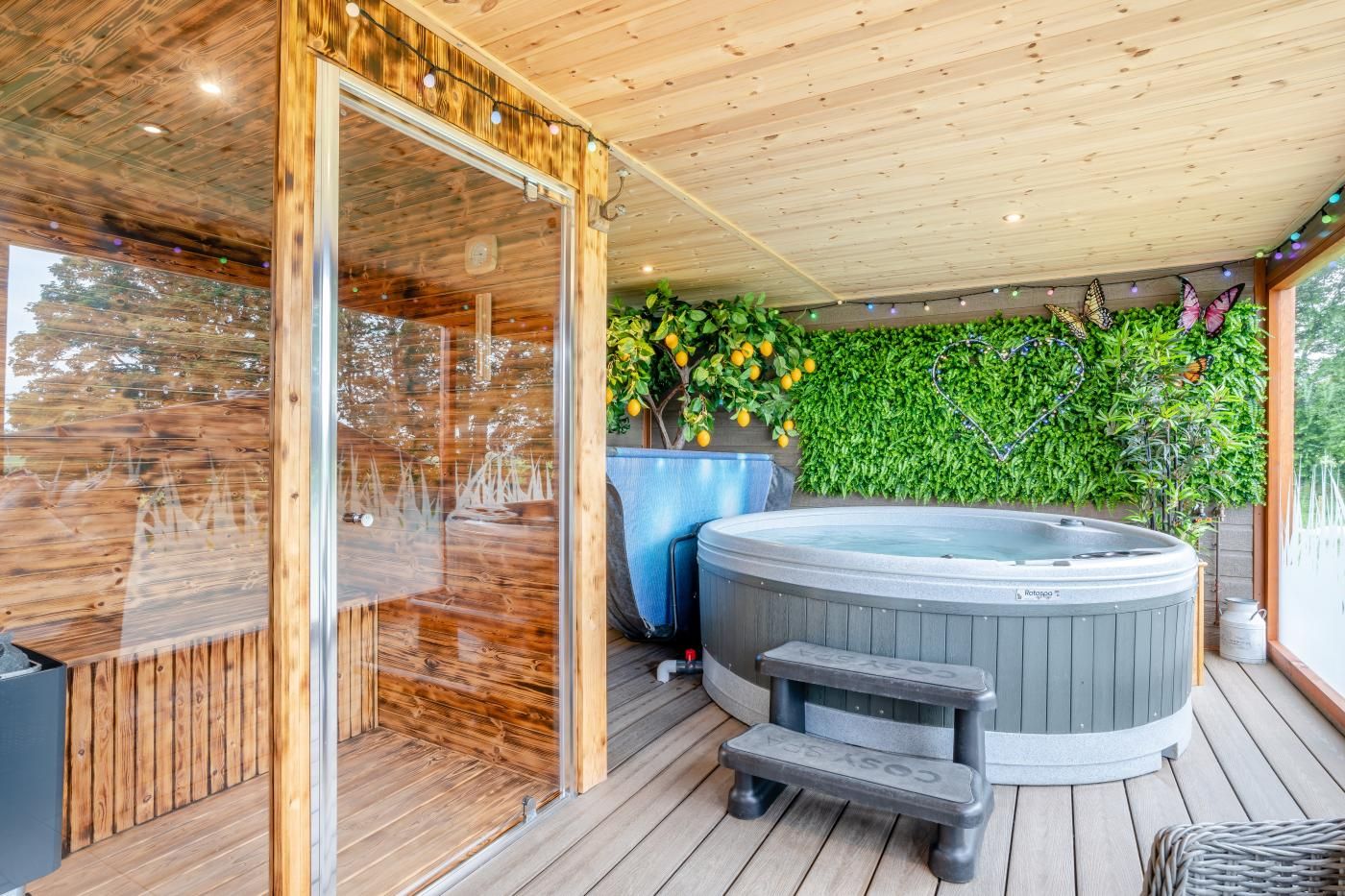 A wooden deck with a hot tub, a glass-enclosed sauna, and a wall of green foliage.