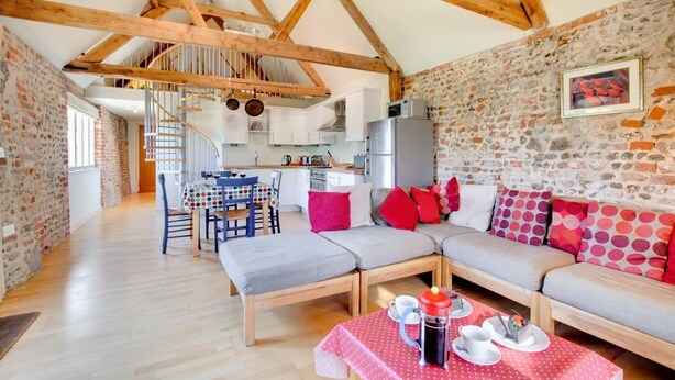 A cozy interior with exposed wooden beams, a stone wall, a dining area with a polka dot tablecloth, and a sofa with colorful pillows. - 2 Bed in Briston (oc-1551) (Briston)