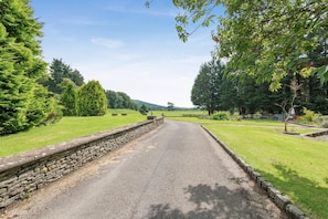 A rural road flanked by stone walls and lush greenery, leading towards distant hills. - 3 Bed in Sedbergh (oc-d32862) (Millthrop)