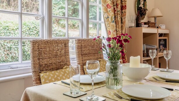 A dining table set with plates, glasses, and a vase of flowers, with wicker chairs and a view of greenery outside. - 2 Bed in Knighton (oc-t32800) (Whitton)