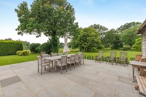 Une terrasse avec une table et des chaises, entourée d’une haie et d’arbres.
