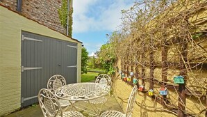 A patio with a round table and chairs, a grey door, and a wall with climbing plants and colorful birdhouses. - 3 Bed in Cromer (oc-1670) (Cromer)