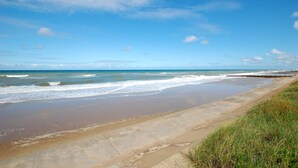 A beach with a concrete walkway along the shore, waves gently crashing onto the sand, and a clear blue sky. - 2 Bed in Bacton (oc-712) (Bacton)