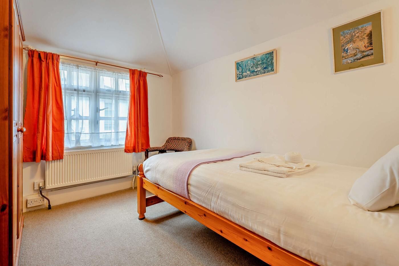 Une chambre avec un lit simple, une chaise, un radiateur et deux tableaux accrochés au mur.
