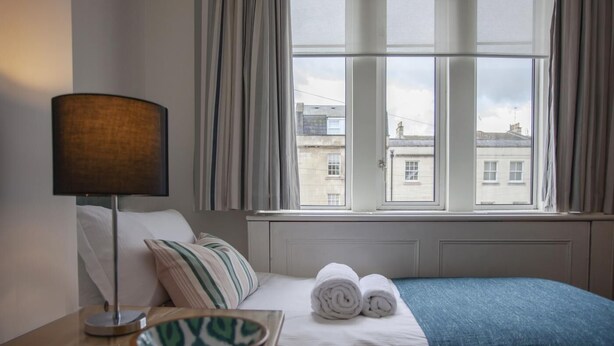 Quarto de hotel com uma cama, uma mesa de cabeceira com abajur, uma tigela e vista para edifícios pela janela.