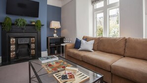 Uma sala de estar com um sofá de couro marrom, uma mesa de centro de vidro com livros e um jogo de tabuleiro, uma lareira com azulejos decorativos, uma TV de tela plana presa na parede e uma janela com cortina estampada.