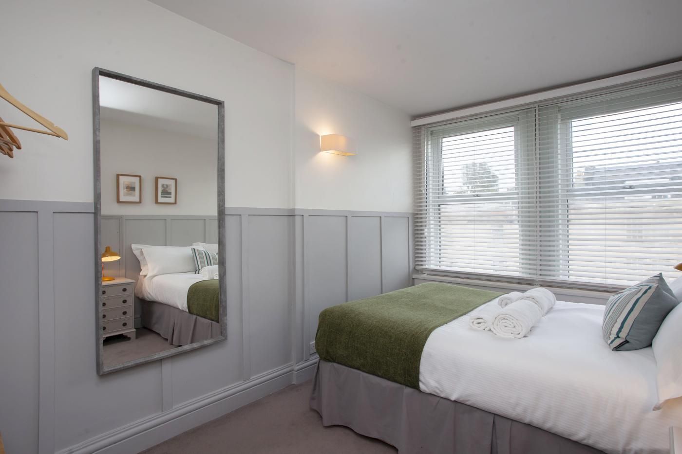 A bedroom with a bed, a bedside table with a lamp, a mirror, and a window with blinds.