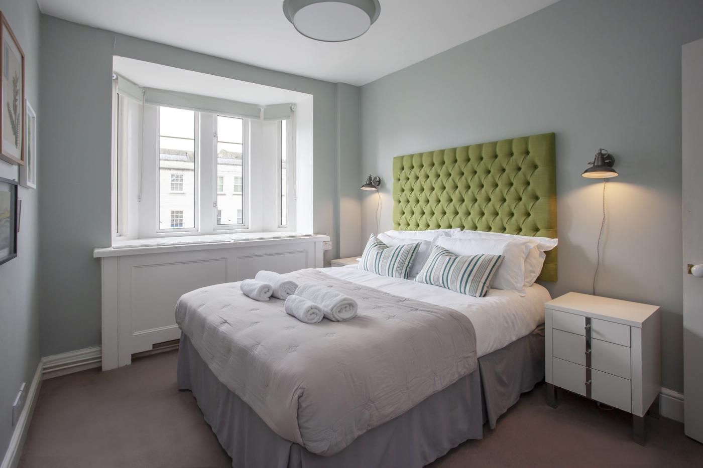 A bedroom with a large bed, a green tufted headboard, a bedside table with a lamp, and a window with white curtains.