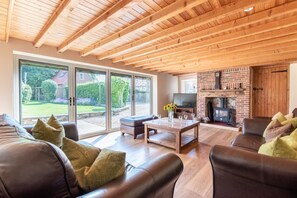 A spacious living room with a wooden ceiling, large sliding glass doors, a brick fireplace, and leather sofas. - 4 Bed in Holme-next-the-Sea (oc-1264) (Holme-next-the-Sea)