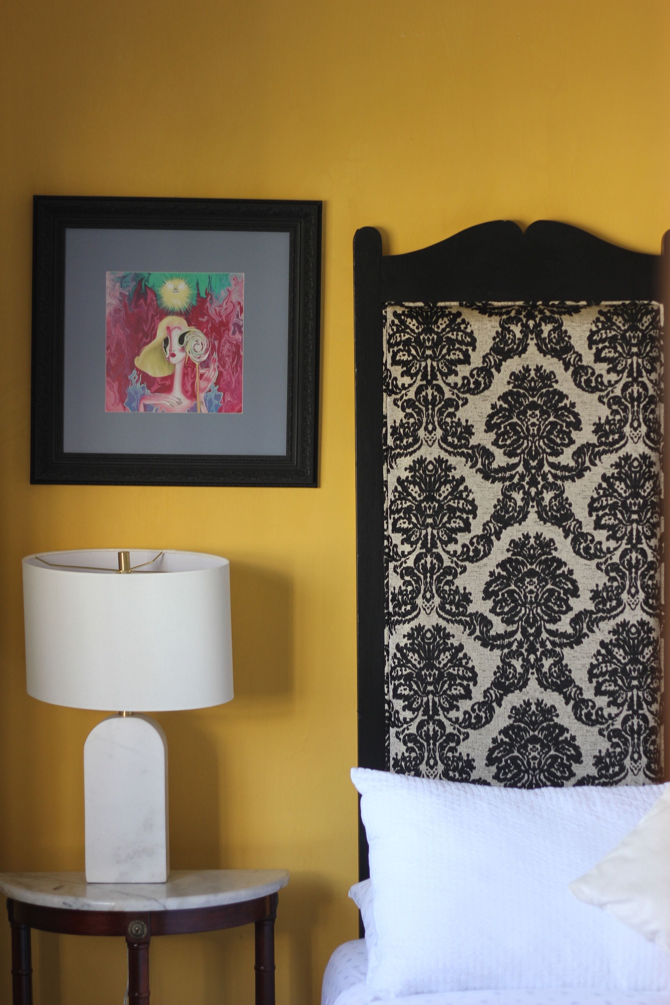 A bed with a headboard, a bedside table with a lamp, and a framed artwork on the wall.