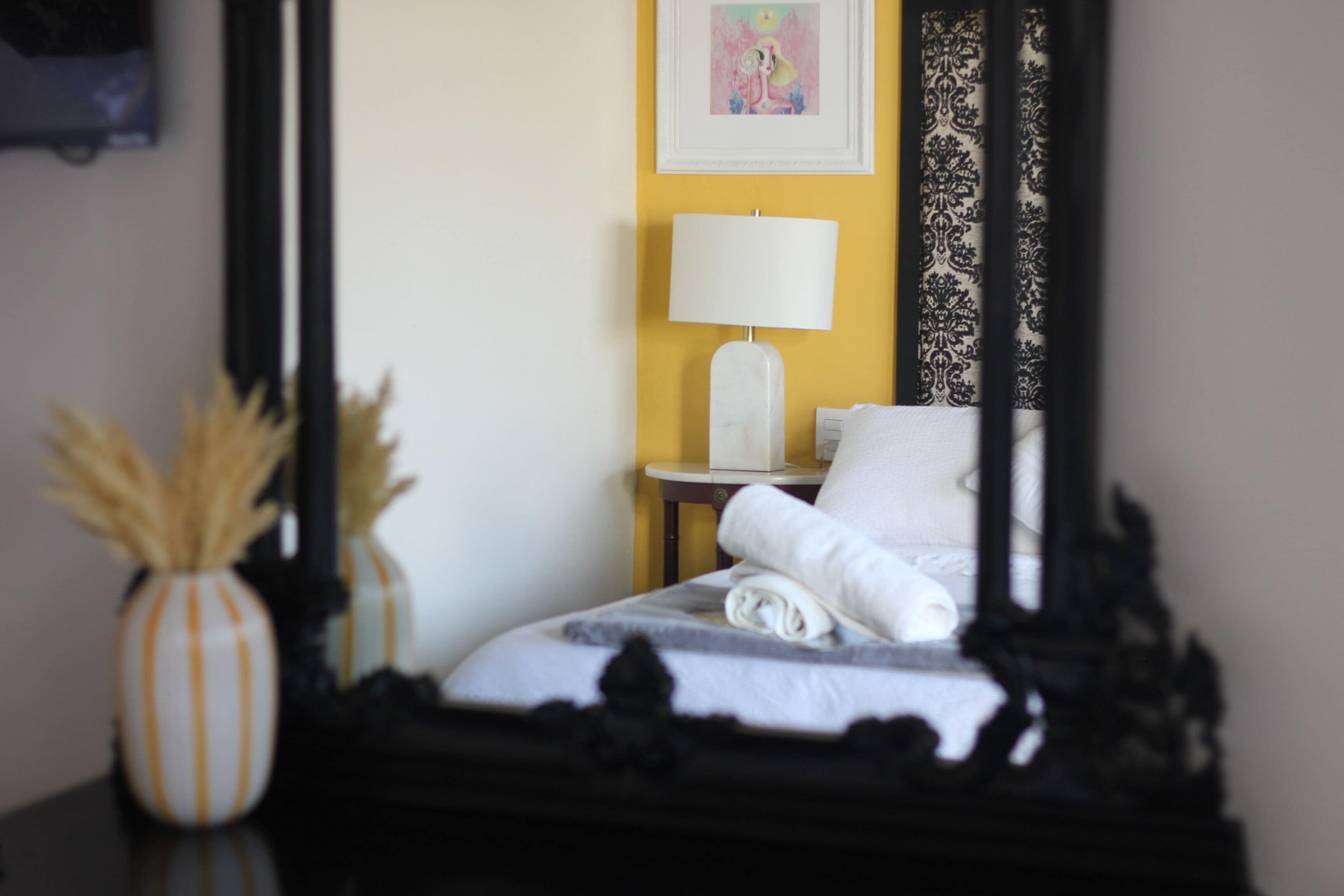 Una habitación de hotel con una cama, una mesita de noche con lámpara, un florero con plantas secas y un cuadro en la pared.