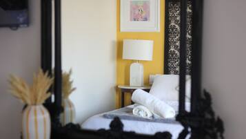 Una habitación de hotel con una cama, una mesita de noche con lámpara, un florero con plantas secas y un cuadro en la pared.