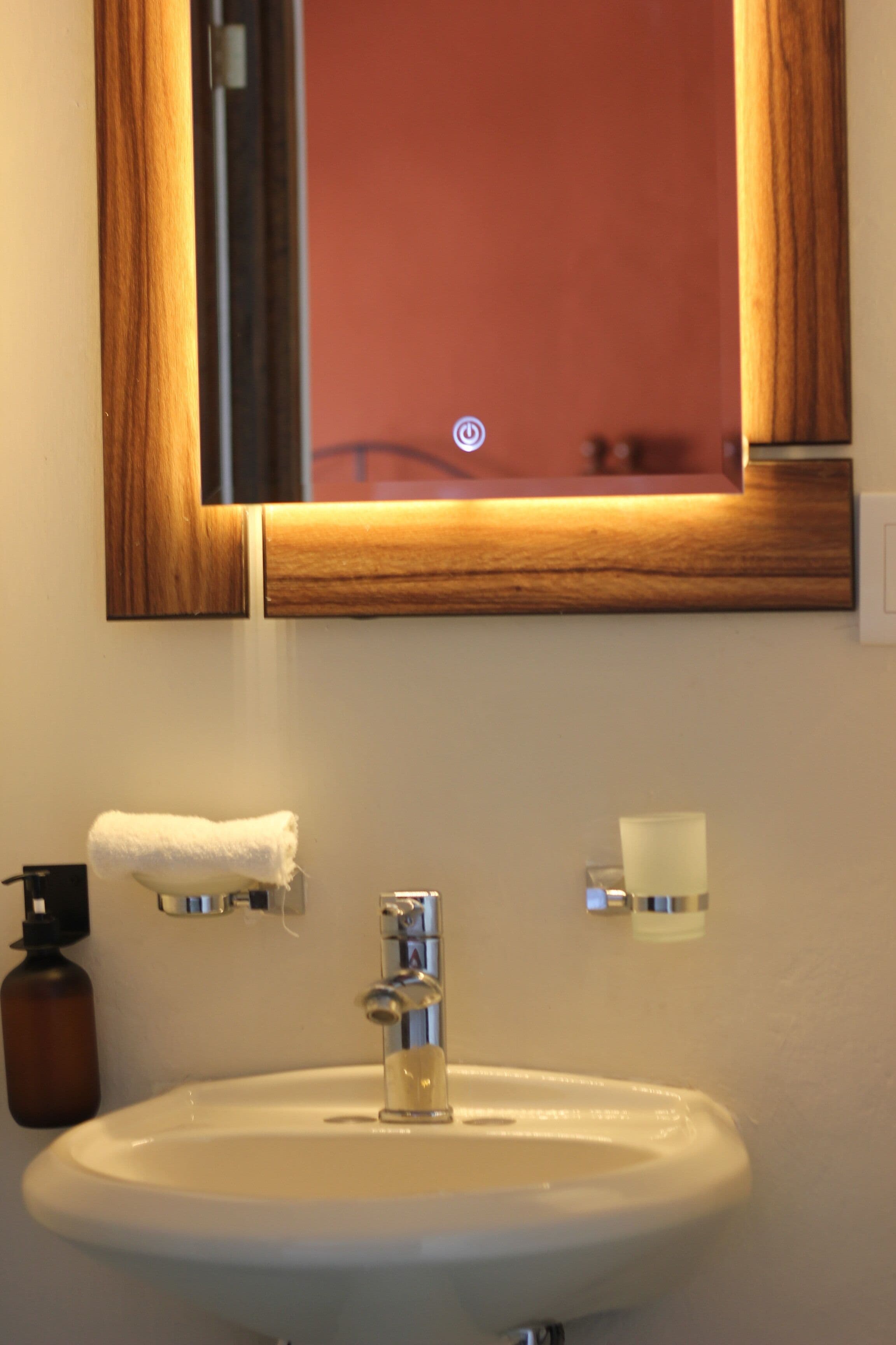 A bathroom with a white sink, a mirror with a wooden frame, a towel hanging on the wall, and a soap dispenser.