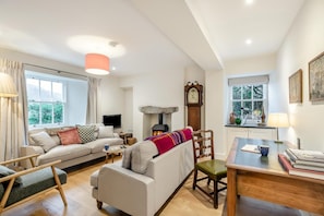 A cozy living room with a sofa, armchair, and a fireplace. There is a dining table with books and a lamp. - 5 Bed in Ulpha (oc-b32731) (Ulpha)