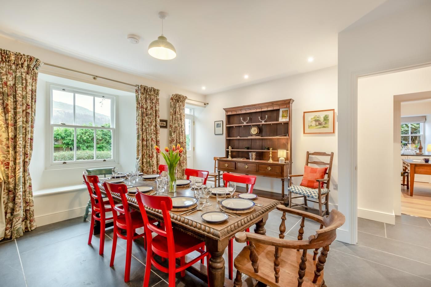 Un coin repas avec une table en bois dressée pour six personnes, entourée de chaises rouges. Une bibliothèque contenant des objets décoratifs est adossée au mur.