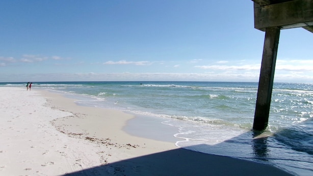 A beach with a concrete pier extending into the water, a clear sky, and footprints on the sand.