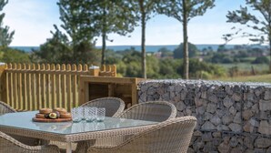 Un espace de restauration en plein air, aménagé avec des meubles en osier et une table en verre, adossé à un mur de pierre et offrant une vue sur les arbres et un paysage lointain.