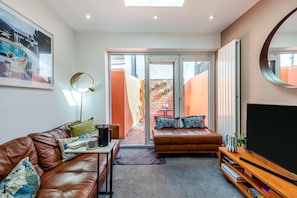 A modern living room with a brown leather sofa, a coffee table, a TV stand with books, and a large window with a view of a brick wall and outdoor furniture.