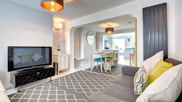 Ein modernes Wohnzimmer mit Flachbildfernseher, einem grauen Sofa mit gemusterten Kissen, einem Essbereich mit Tisch und Stühlen sowie einer Küche im Hintergrund.