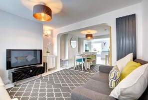 Un salon moderne avec un téléviseur à écran plat, un canapé gris agrémenté de coussins à motifs, un coin repas avec une table et des chaises, et une cuisine en arrière-plan.
