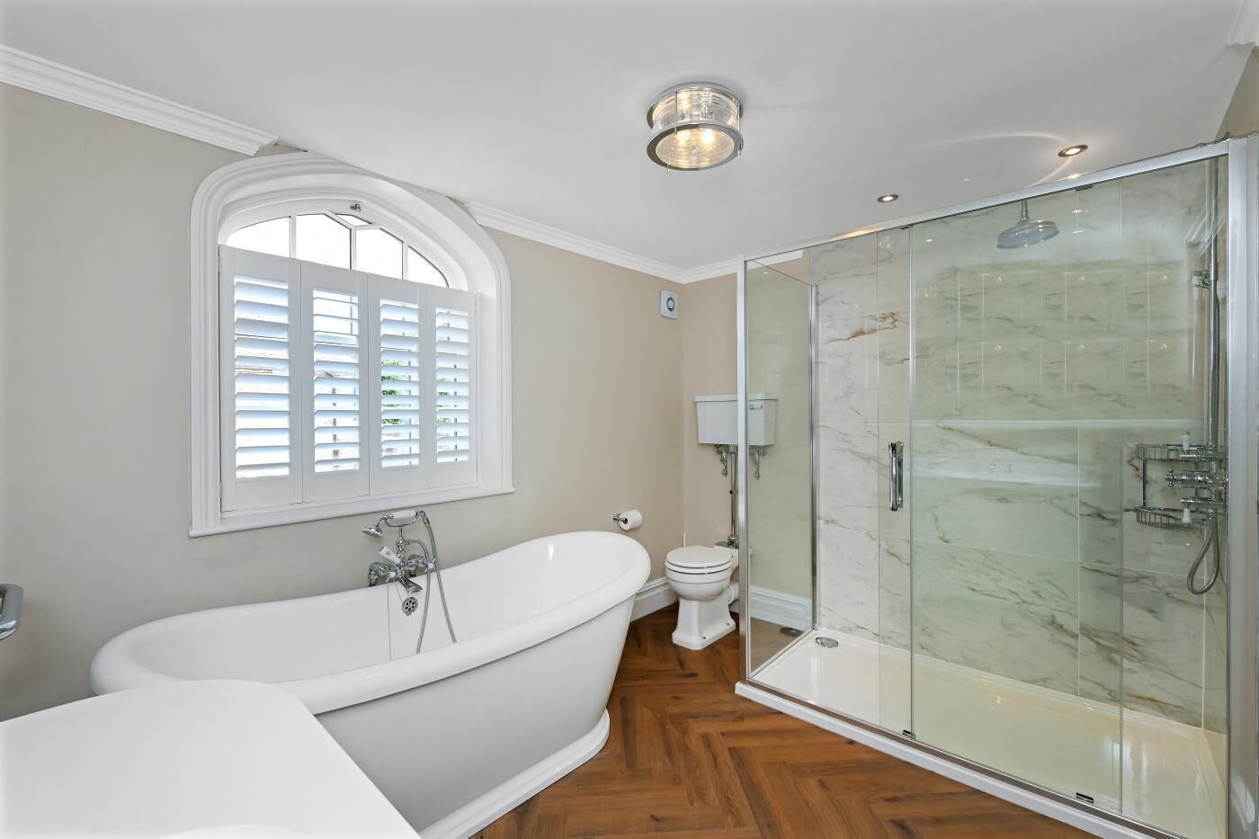 A bathroom with a freestanding bathtub, a glass-enclosed shower, a toilet, and a window with shutters.