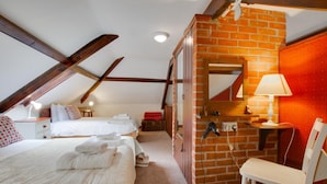 A cozy attic bedroom with a sloped ceiling, a brick wall, and a wooden beam structure. There is a bed with white linens and a chair with a lamp. - 1 Bed in Banningham (oc-564) (Banningham)