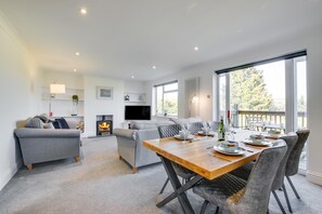 A modern living room with a fireplace, a dining area with a wooden table set for four, and a balcony with outdoor seating. - 3 Bed in Ovingdean (oc-bbwand) (Brighton)