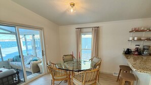 Uma área de refeições com mesa de vidro e cadeiras de vime, uma cozinha com bancada de granito e vista para o mar através de amplas janelas.