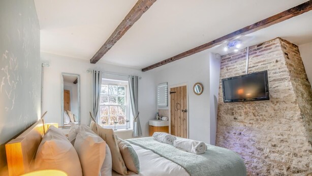 Et soveværelse med en seng, en sengetavlelampe, et vindue med gardiner, en trædør, et spejl, et fjernsyn ophængt på en stenvæg og et lille bord med en lampe.