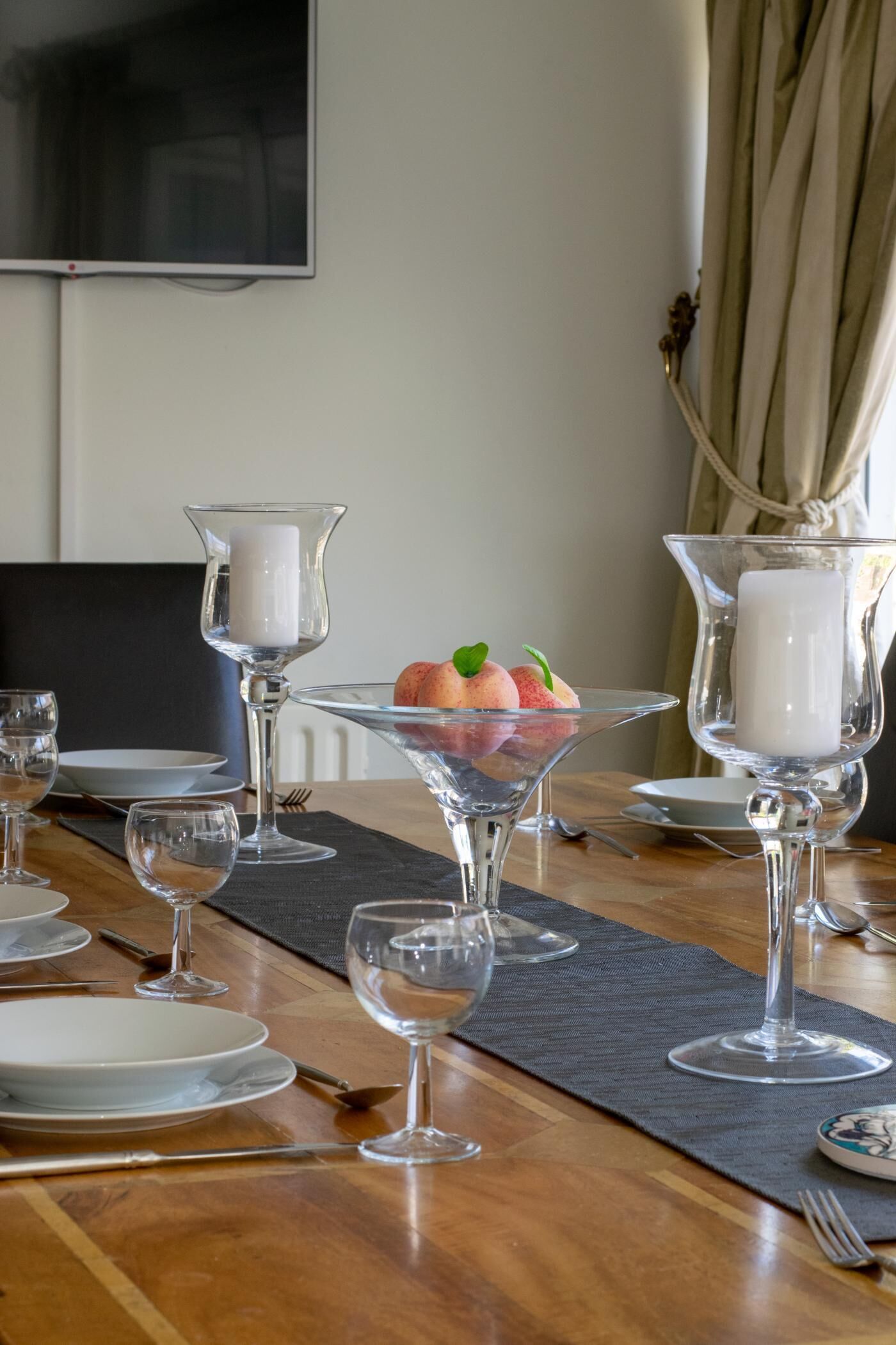 Mesa posta com copos de vidro transparente, pratos e um arranjo de frutas.