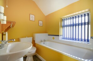 Ein Badezimmer mit weißem Waschbecken, Toilette und Badewanne. An der Wand hängen zwei Bilder und ein Fenster mit blauen Jalousien.