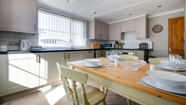A modern kitchen with white cabinets, a wooden dining table set with plates and glasses, and a window with blinds. - 2 Bed in Holt (oc-1449) (Holt)