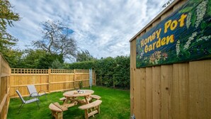 Un patio trasero con valla de madera, mesa de picnic y sillas. En la valla hay un letrero que dice 'Honey Pot Garden'.