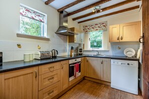 A kitchen with wooden cabinets, a dishwasher, a stove, and a window with floral curtains. - 2 Bed in Rolvenden (oc-tn563) (Rolvenden)