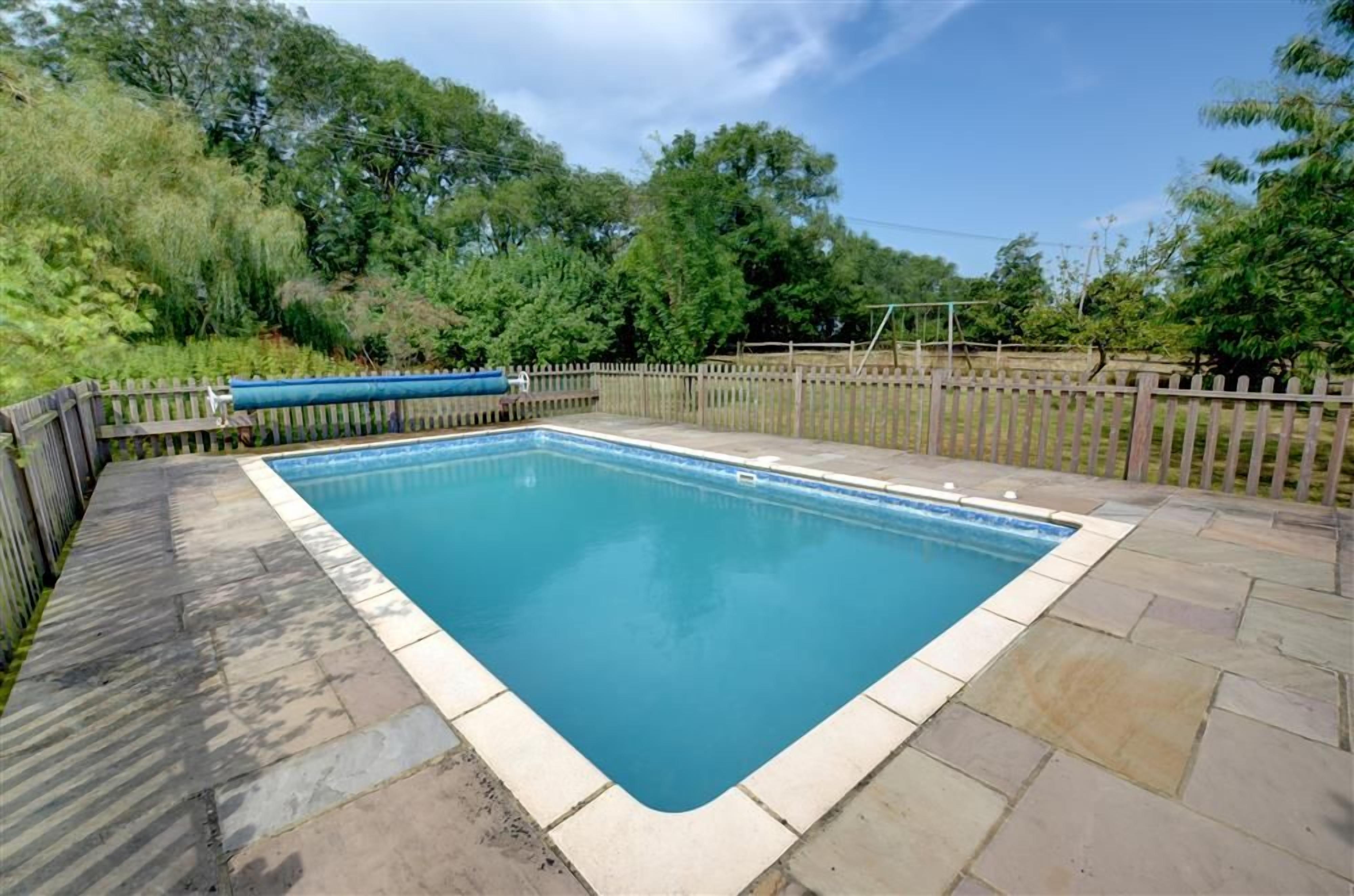 A rectangular swimming pool with a tiled edge, surrounded by a wooden fence and a stone patio.