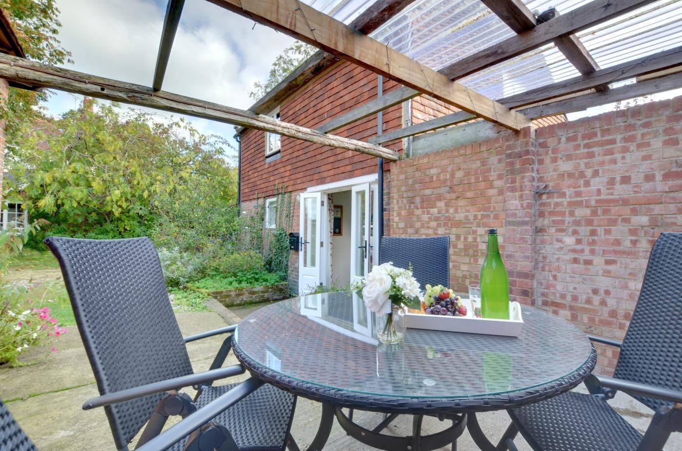 A covered patio with a round glass table, four wicker chairs, a brick wall, and a wooden beam structure.