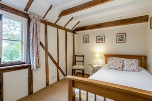 A bedroom with a wooden ceiling, a bed with floral bedding, a chair, a bedside table with a lamp, and a window with curtains. - 2 Bed in Rolvenden (oc-tn563) (Rolvenden)