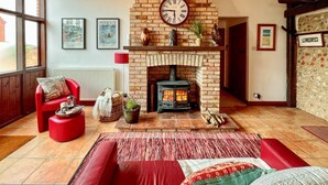 A cozy living room with a brick fireplace, a red sofa, and a red armchair. There are framed pictures on the wall and a window with a view outside. - 3 Bed in Holt (oc-1331) (Bodham)