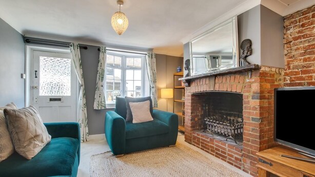 A living room with a brick fireplace, a teal sofa, a green armchair, and a television. - 4 Bed in Rottingdean (oc-bbrott) (Brighton)