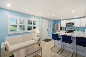 A compact kitchen with white cabinets, a microwave, and a refrigerator. There is a dining area with blue chairs and a table. A beige sofa is placed near a window with blinds. - ⭐Charming Triplex | Close to Beach | Pet Friendly | The Starfish Estate⭐ (Fort Myers Beach)