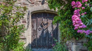 A rustic stone building with a weathered wooden door, a heart-shaped decoration, and lush greenery.