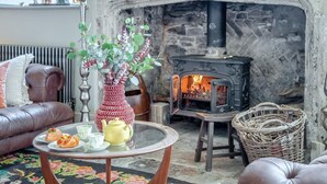 A cozy living room with a fireplace, leather sofa, a coffee table with pastries, and a vase with flowers.