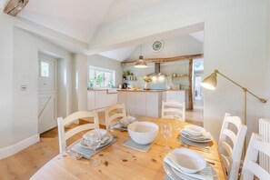 A dining area with a wooden table set for four, white chairs, and a kitchen in the background. - 3 Bed in Sedlescombe (oc-c32764) (Sedlescombe)