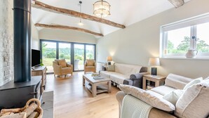A cozy living room with a fireplace, wicker furniture, and a large window offering a view of the outdoors. - 3 Bed in Sedlescombe (oc-c32764) (Sedlescombe)