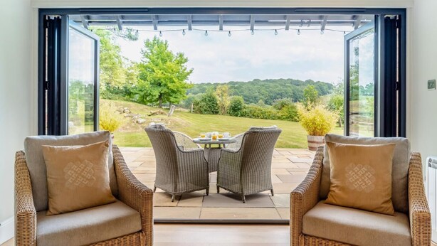 A room with a large sliding glass door leading to a patio with wicker furniture and a table, overlooking a green landscape. - 3 Bed in Sedlescombe (oc-c32764) (Sedlescombe)