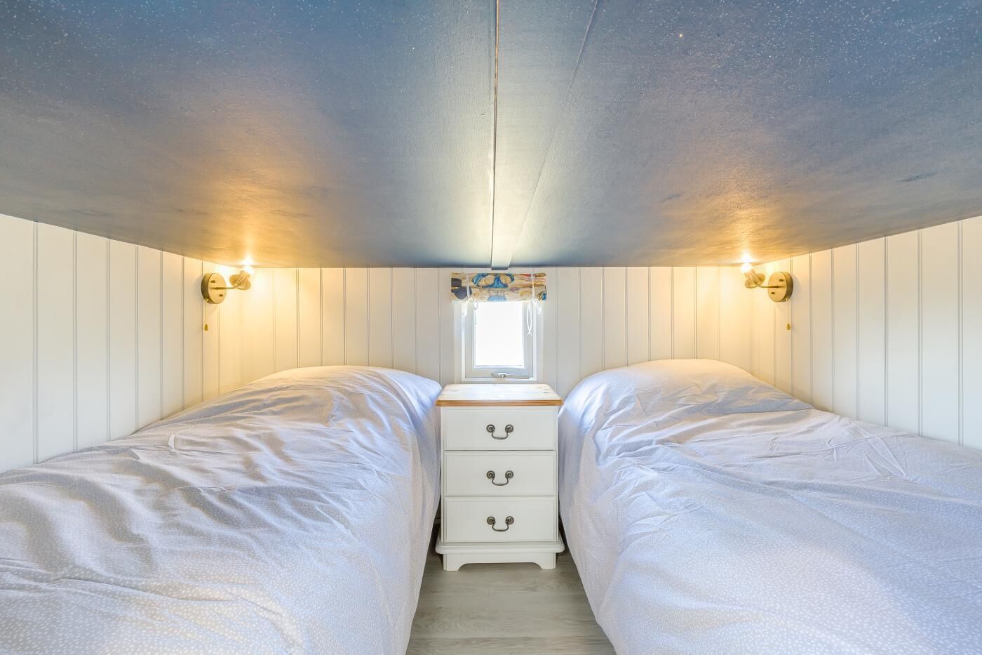 Quarto com duas camas, uma janela pequena e luminárias na parede.