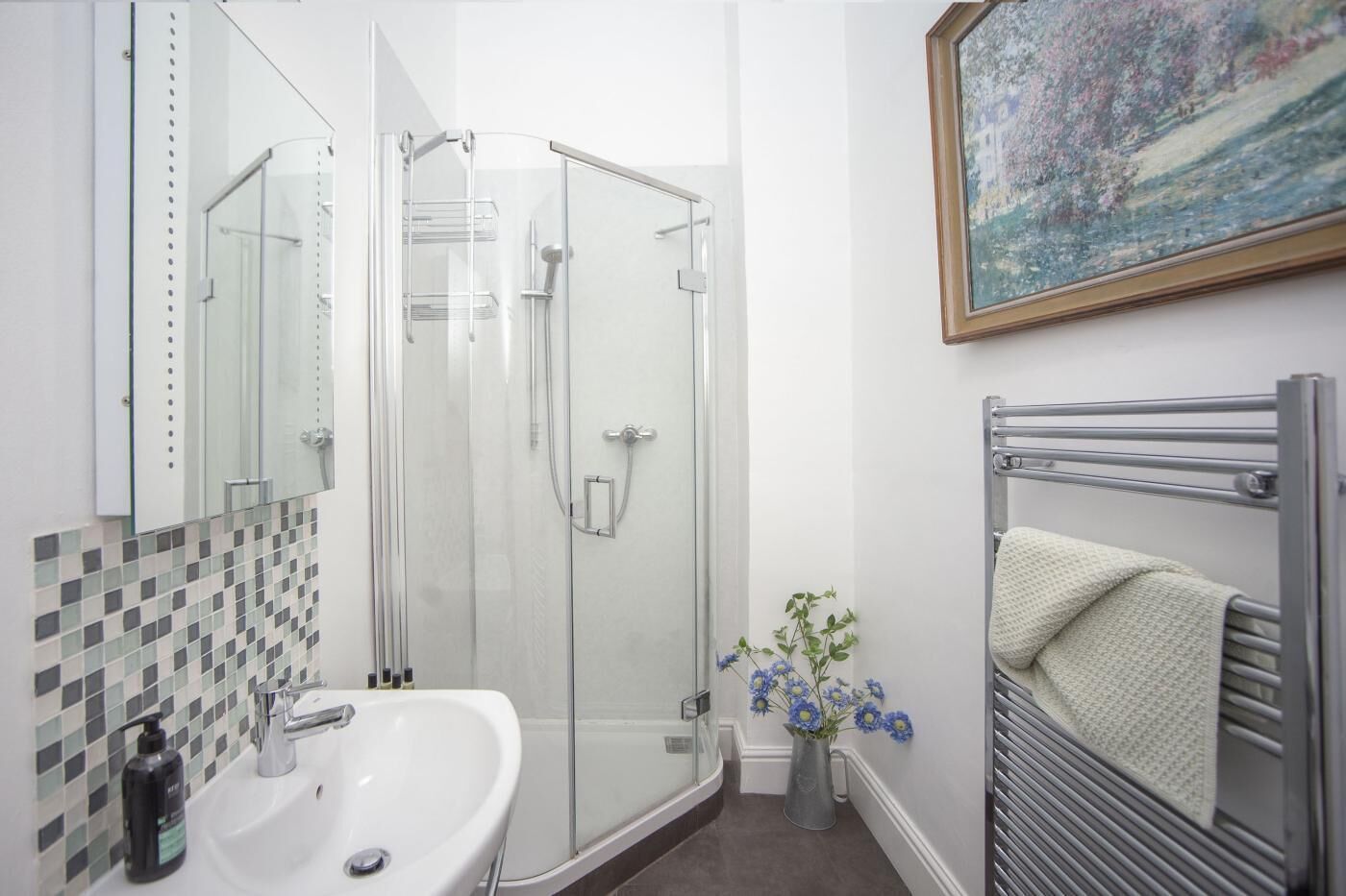 A bathroom with a glass shower enclosure, a white sink, a towel rack with a towel, and a painting on the wall.