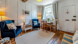 A cozy living room with a blue armchair, a white side table, a dining table with chairs, and a large window with curtains. - 2 Bed in Rye (oc-rh1045) (Rye)