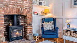 A cozy room with a brick fireplace, a blue armchair, a small table with a lamp, and a wicker basket. - 2 Bed in Rye (oc-rh1045) (Rye)